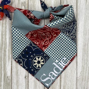 May include: A triangular pet bandana featuring a patchwork design of red, white, and blue patterns including gingham, paisley, and floral motifs. The name Sadie is embroidered in white script on the lower corner. A small patriotic pinwheel is attached.