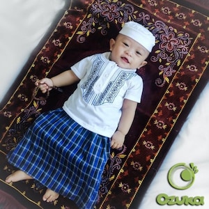 May include: A baby boy wearing a white shirt with a geometric pattern, a blue and white plaid skirt, and a white skullcap. He is lying on a red prayer rug with gold and brown floral designs.