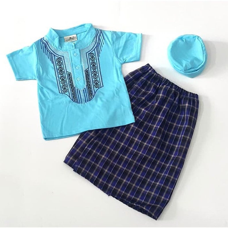May include: A light blue short-sleeved shirt with an embroidered design, a blue and black plaid skirt, and a light blue hat. The shirt has a button closure at the neck.
