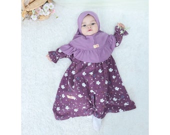 May include: A baby girl wearing a purple floral print dress with a matching purple hijab. The dress has long sleeves and a ruffled neckline. The baby is lying on a white fluffy surface.