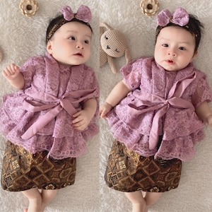 May include: A baby wearing a purple lace dress with a brown patterned skirt and a purple bow headband. The baby is sitting on a white fuzzy surface.