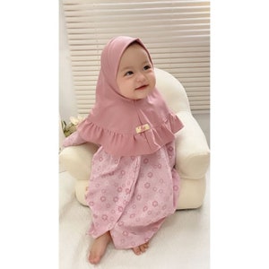 May include: A pink baby dress with a floral pattern and a matching pink ruffled hijab. The hijab has a small yellow tag with the word "life" on it.