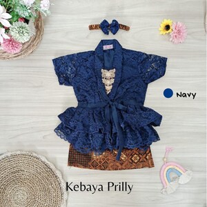 May include: A navy blue lace kebaya with a ruffled hem and a matching headband. The kebaya is paired with a brown and gold batik skirt. The text "Kebaya Prilly" is visible below the skirt.