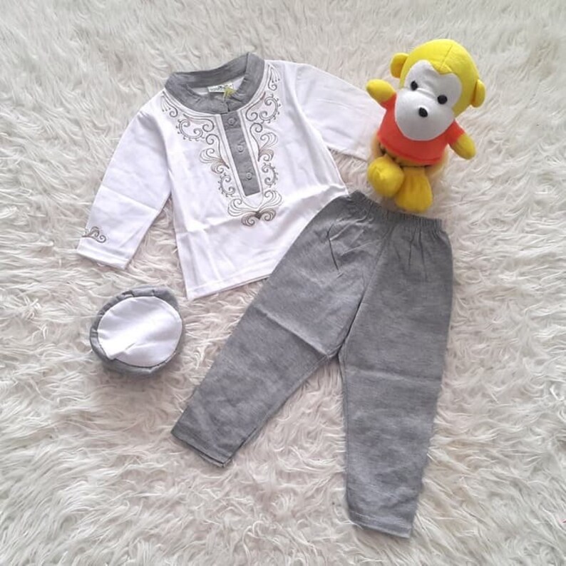 May include: A white long-sleeved shirt with a gold embroidered design, gray pants, and a white cap. The shirt has a button closure at the neck.