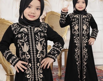 May include: Two girls wearing black abayas with gold embroidery. The abayas have long sleeves and a long skirt. The girls are wearing headscarves.