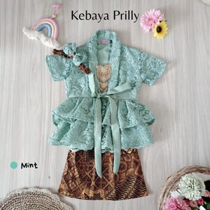 May include: Mint green lace kebaya with a ruffled skirt and a matching headband. The kebaya has a sash tied at the waist and a gold sequin embellishment. The skirt is brown and white with a batik pattern.