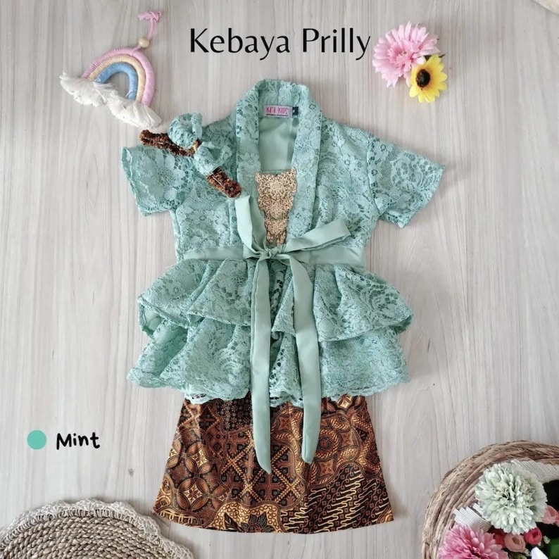 May include: Mint green lace kebaya with a ruffled skirt and a matching headband. The kebaya has a sash tied at the waist and a gold sequin embellishment. The skirt is brown and white with a batik pattern.