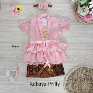 May include: A pink lace kebaya with a ruffled skirt and a matching headband. The kebaya has a gold embroidered design on the front and a pink ribbon tied at the waist. The skirt is a brown and gold batik print. The text "Kebaya Prilly" is written below the skirt.