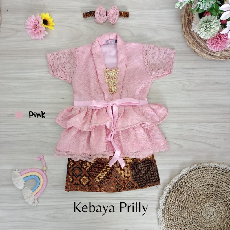 May include: A pink lace kebaya with a ruffled skirt and a matching headband. The kebaya has a gold embroidered design on the front and a pink ribbon tied at the waist. The skirt is a brown and gold batik print. The text "Kebaya Prilly" is written below the skirt.