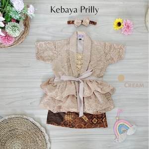 May include: A cream-colored lace kebaya with a ruffled skirt and a matching sash. The kebaya has a gold embroidered design and a brown batik skirt. The outfit is accessorized with a cream-colored headband with a bow.