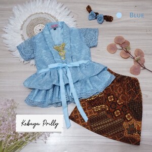 May include: A light blue lace kebaya with a gold embellishment and a matching blue ribbon. The kebaya is paired with a brown batik skirt. The text "Kebaya Prilly" is visible in the image.
