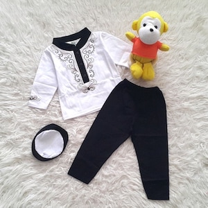 May include: A white long-sleeved shirt with a brown embroidered design, black pants, and a white and black hat. A yellow stuffed monkey is also in the image.
