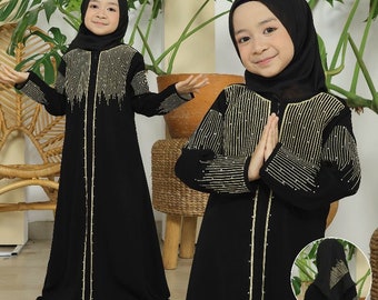 May include: Black abaya dress with gold vertical embellishments on the bodice and sleeves. The dress features a matching black hijab. The dress has a vertical gold trim down the front. The text "Kerudung belakang" is visible in the image.