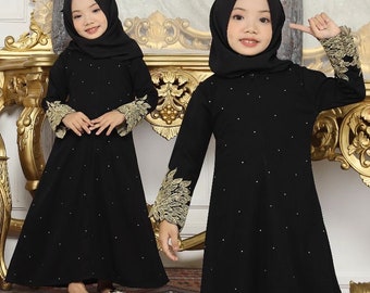 May include: Two girls wearing black abayas with long sleeves and gold embroidery on the cuffs. The abayas are adorned with small, scattered white embellishments. Both girls are wearing black hijabs and are standing in front of a gold-framed mirror.