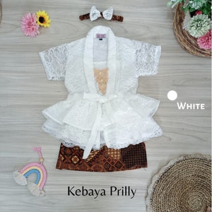 May include: White lace kebaya with a ruffled skirt and a matching headband. The kebaya has a bow at the waist and a gold beaded embellishment on the front. The skirt is brown and has a batik pattern. The text "Kebaya Prilly" is written below the image.