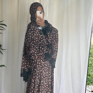 May include: A floral print dress and head covering in a dark color palette with small pink and white flowers. The dress features black lace trim on the sleeves and neckline. The outfit is worn by a person.
