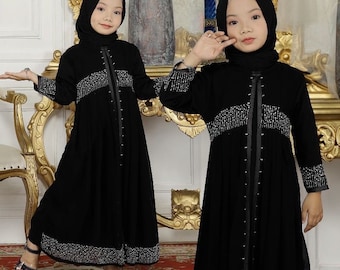 May include: Two young girls wearing black abayas with silver bead embellishments. The abayas have long sleeves and a long skirt. The girls are wearing head scarves and crowns.