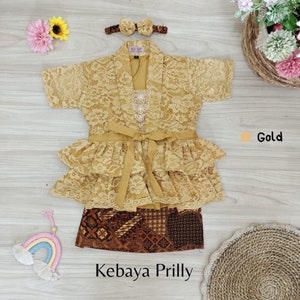 May include: A gold lace kebaya with a ruffled skirt and a matching headband. The kebaya has a brown batik skirt underneath. The text "Kebaya Prilly" is written below the image.