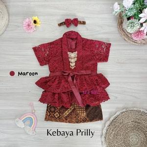 May include: A maroon lace kebaya with a ruffled skirt and a gold necklace. The kebaya is tied with a maroon ribbon and has a matching headband with a bow. The text "Kebaya Prilly" is written below the image.