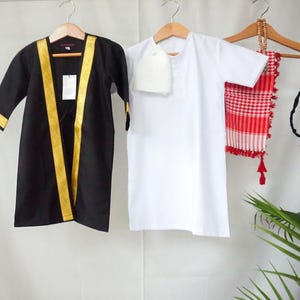 May include: Two traditional Arab garments hang on wooden hangers. One is a black robe with gold trim, the other is a white shirt with a matching head covering. A red and white checkered scarf and a black tassel bag are also displayed.