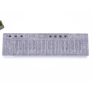 Puede incluir: Un teclado de fieltro gris con teclas y botones negros. El teclado es una versión en miniatura de un teclado real.