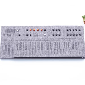 Puede incluir: Un sintetizador gris con teclado, perillas y luces naranjas. El instrumento está hecho de un material similar al fieltro y se encuentra sobre una superficie blanca. Una pequeña planta en maceta está en la esquina superior derecha.