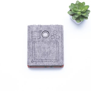 May include: A gray felt notebook with a black outline of a camera on the front. The notebook has a small circle in the center of the camera outline and three smaller circles on the right side of the camera outline.