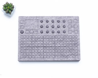 May include: A gray felt pad with a grid of squares and a row of round buttons. The pad is designed for use with a digital music controller.