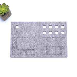 May include: A gray felt desk mat with a grid pattern and cutouts for organizing small items. The mat has a rectangular shape and is designed for use on a desk.