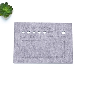 Peut inclure: Un organisateur en feutre gris avec plusieurs trous et sections rectangulaires. Une petite plante succulente verte dans un pot blanc est dans le coin supérieur gauche. L'organisateur est sur fond blanc.