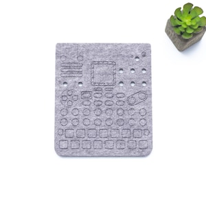 Op de afbeelding: Een grijs viltpad met een raster van cirkels en vierkanten, ontworpen voor gebruik met elektronische muziekapparatuur. Het pad heeft een klein rechthoekig gebied in het midden en twee kleine cirkels aan de rechterkant.