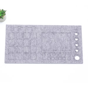 Puede incluir: Una alfombrilla de teclado de fieltro gris con contornos de un teclado, una pantalla y botones. La alfombrilla está diseñada para parecerse a un controlador de producción musical.