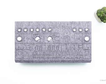 May include: A rectangular gray felt cover for a music synthesizer, featuring laser-cut circular openings for knobs and rectangular slots for sliders. The surface includes engraved outlines of a control panel layout and a small smiley face icon.