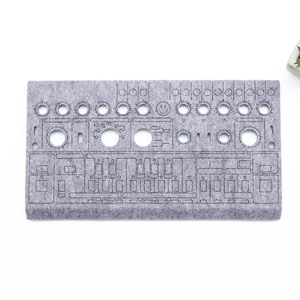 Könnte beinhalten: Ein graues Filzpaneel mit einem detaillierten Ausschnittdesign, das einem Synthesizer-Bedienfeld ähnelt. Das Panel hat verschiedene kreisförmige und rechteckige Ausschnitte, mit einer kleinen Pflanze in einem weißen Topf in der oberen rechten Ecke. Der Text "WaveTable 2026" befindet sich in der unteren rechten Ecke.