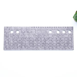 Könnte beinhalten: Ein graues Filz-Keyboard-förmiges Objekt mit Tastenkonturen und kreisförmigen Löchern. Eine kleine grüne Pflanze mit runden Blättern befindet sich in der oberen rechten Ecke. Das Objekt befindet sich auf weißem Hintergrund.