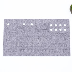 Peut inclure: Un tapis de bureau en feutre gris avec une disposition de clavier et des trous pour la gestion des câbles. Le tapis a une petite zone rectangulaire avec un motif hachuré.