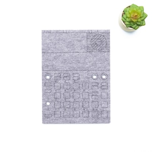 Puede incluir: Un panel de sintetizador de fieltro gris con una cuadrícula de botones y una sección superior con un diseño circular. Una pequeña suculenta en una maceta blanca se encuentra en la esquina superior derecha. El texto "WaveTable 2025" está en la esquina inferior derecha.