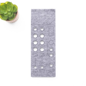 May include: A gray felt panel with various cut-out circles and rectangular shapes, resembling a control panel. The panel has a stitched border and is placed on a white surface. A small potted succulent is in the upper left corner.