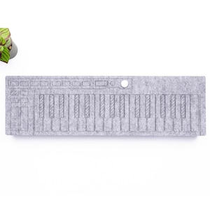 Puede incluir: Un organizador gris en forma de teclado de fieltro con un botón blanco y una pequeña planta verde en una maceta. El teclado tiene una serie de teclas negras y grises y botones de control. Copyright WaveTable 2025.