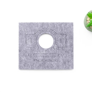 May include: A gray felt WaveTable component with a circular cutout and etched design. The design includes a central circle, a series of lines, and a row of six small circles. A small potted succulent is in the upper right corner.