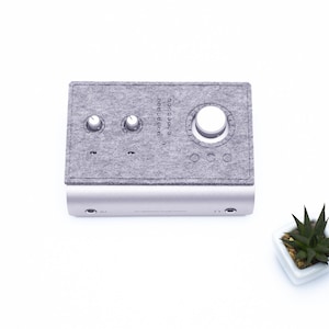 Puede incluir: Una interfaz de audio plateada y gris con tres botones y una cubierta de tela gris. La interfaz tiene dos conectores de audio en la parte delantera.