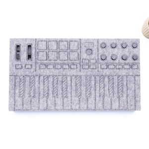 Op de afbeelding: Een grijze vilten synthesizer met een toetsenbord, knoppen en pads. Het ontwerp is in het vilt geëtst. Een kleine vetplant in pot staat in de rechterbovenhoek. De copyrightvermelding "WaveTable 2025" staat in de rechteronderhoek.