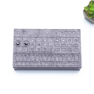 Op de afbeelding: Een grijze vilt drummachine met een zwarte omtrek van de knoppen en knoppen. De drummachine staat op een witte achtergrond.