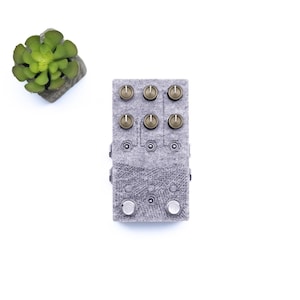 Puede incluir: Un pedal de música electrónica gris y blanco con seis botones y dos botones. Los botones son de color dorado. El pedal tiene una superficie texturizada.