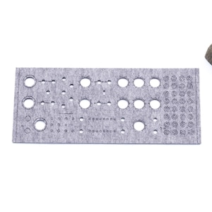 Puede incluir: Un panel de fieltro gris con varios agujeros circulares y pequeños de diferentes tamaños, y un patrón de pequeños círculos en el lado derecho. Una pequeña planta suculenta en maceta se encuentra en la parte superior derecha. El texto "WaveTable 2025" está en la esquina inferior derecha.