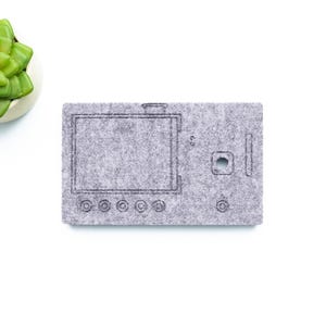 May include: A grey felt WaveTable 2026 panel with a detailed design, featuring a screen outline, buttons, and a port. A small succulent in a white pot is in the upper left corner. The panel is on a white background.