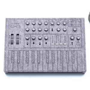 May include: A grey felt-like mat with a design of a synthesiser keyboard. The mat has a series of knobs, sliders, and keys.