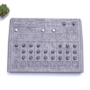 Könnte beinhalten: Eine graue Filzmatte mit dem Design eines Drumcomputers. Die Matte hat eine Vielzahl von Knöpfen, Drehknöpfen und Schiebereglern.