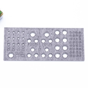 Puede incluir: Un panel de fieltro gris con varios recortes circulares y rectangulares, probablemente para un sintetizador modular o un proyecto electrónico. Una pequeña planta suculenta en una maceta cuadrada blanca está en la esquina superior izquierda.