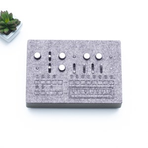 May include: A grey felt music sampler with white knobs and buttons. The sampler has a grid of squares and a row of buttons labelled "PLAY" and "STOP".